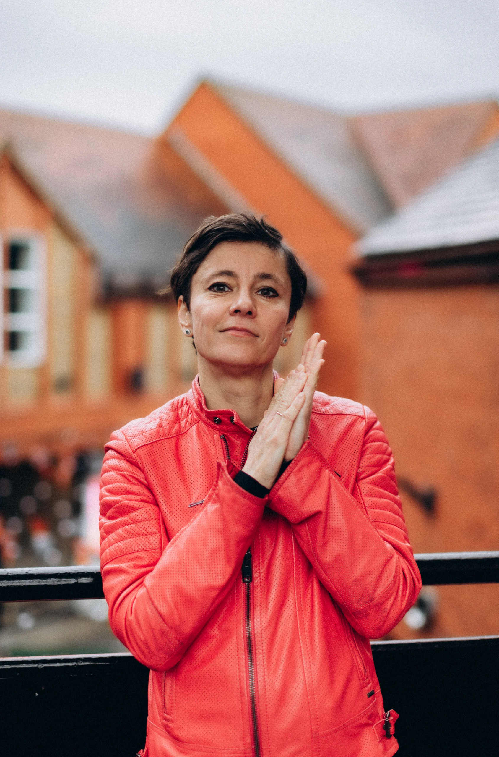 Irina Dmitrieva standing outdoors in a bright red jacket, hands pressed together, with a warm brick building in the background