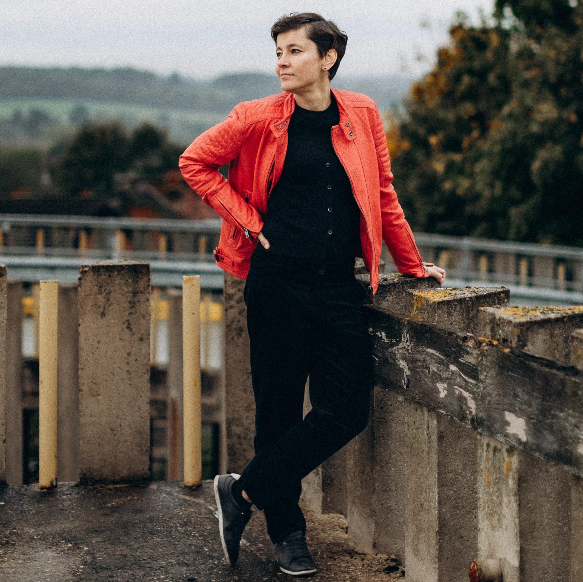 Irina Dmitrieva wearing an orange jacket, standing on a rooftop or elevated terrace, looking away from the camera towards an open landscape.