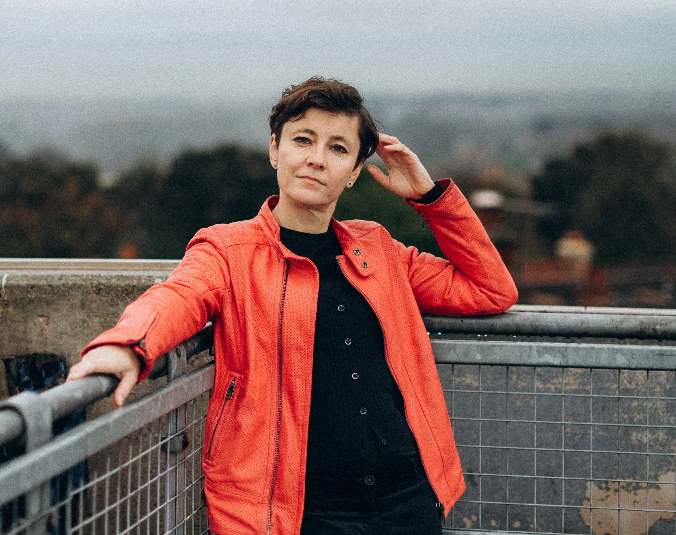 Irina Dmitrieva in an orange jacket standing on a rooftop terrace, looking out over a wide open landscape.