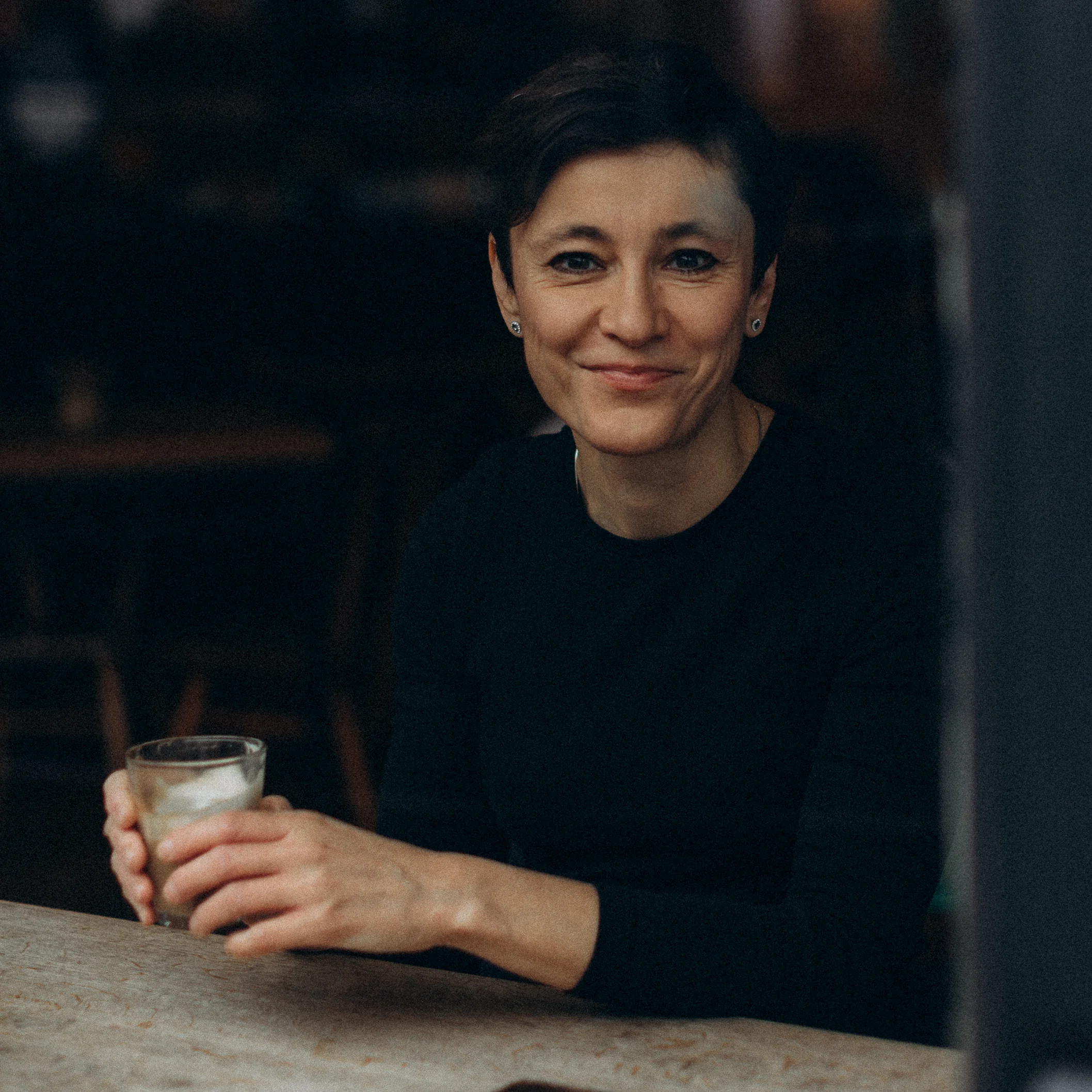 Irina Dmitrieva seated at a cafe table, holding a drink, smiling warmly at the camera in a relaxed indoor setting.